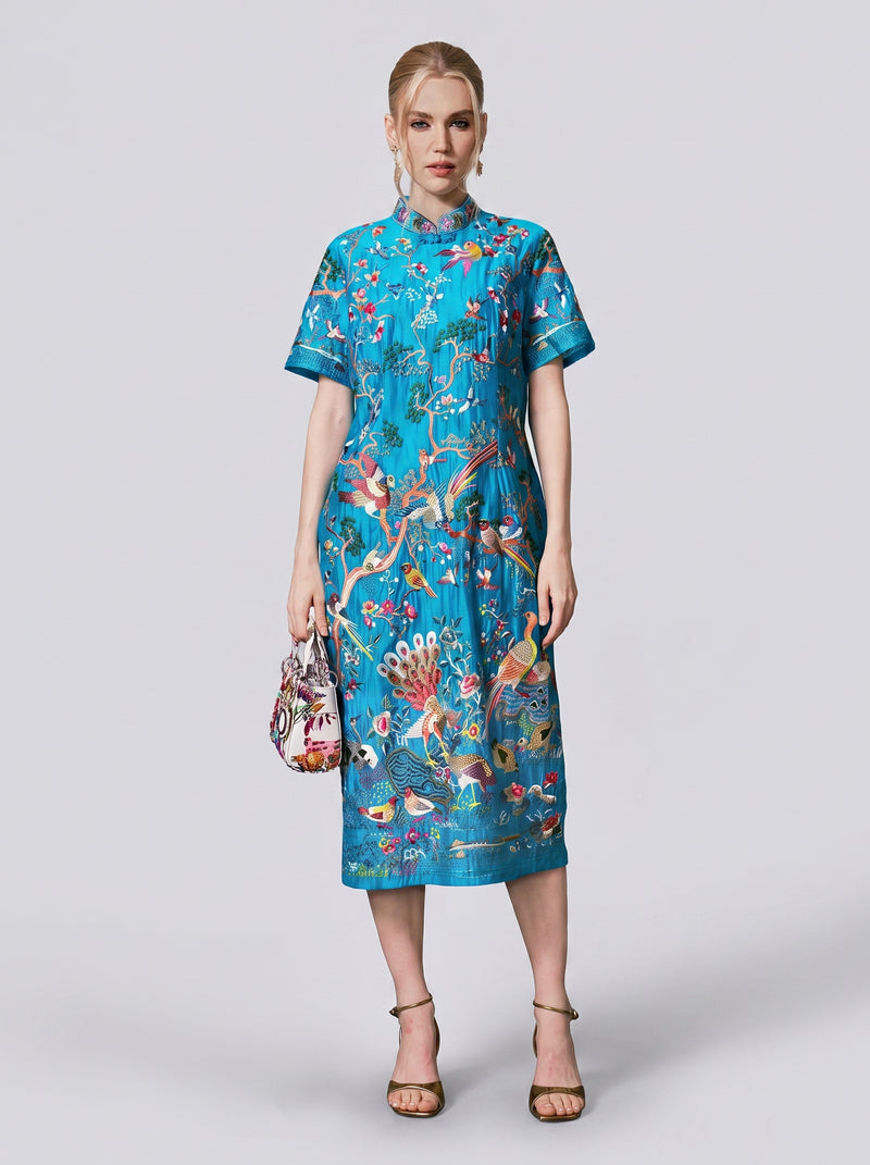 A woman stands facing forward wearing the Social Queen Allover Bird-Embroidered Dress, a turquoise, knee-length piece adorned with colorful bird and floral patterns. She holds a small handbag and wears strappy sandals.
