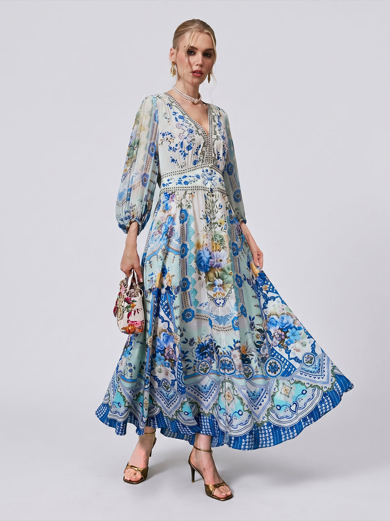 A woman stands against a plain background wearing the Social Queen Floral Print Mulberry Silk Dress, featuring a long, flowy silhouette with blue floral and geometric patterns. She holds a small handbag and wears heeled sandals adorned with subtle rhinestone accents.