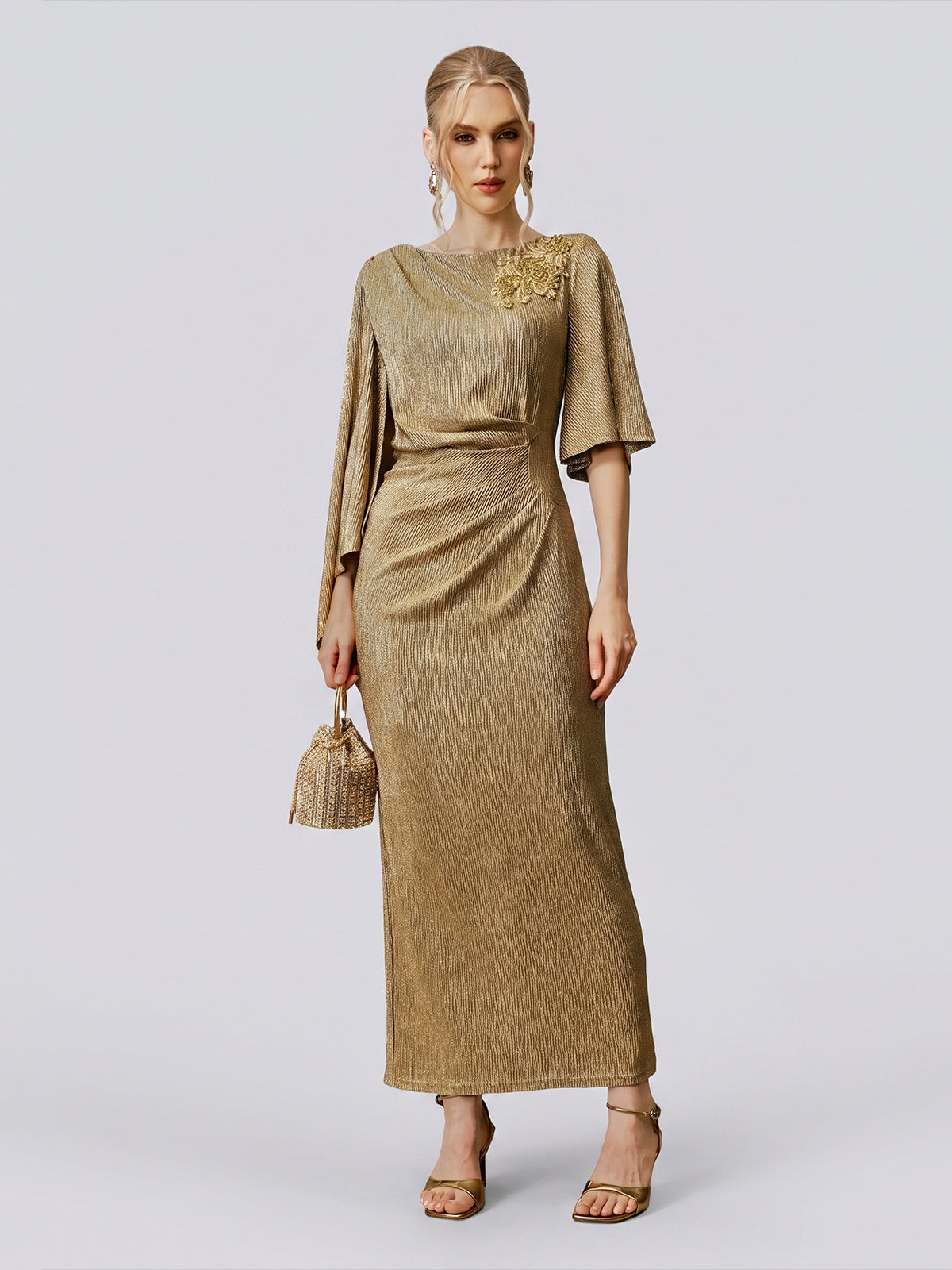 A woman stands against a plain background wearing the Floral Beaded Knit Dress by Social Queen—a gold, floor-length evening gown featuring intricate beading and draped sleeves. She accessorizes with a small handbag, matching heels, and coordinating earrings.