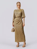 A woman stands against a plain background wearing the Floral Beaded Knit Dress by Social Queen—a gold, floor-length evening gown featuring intricate beading and draped sleeves. She accessorizes with a small handbag, matching heels, and coordinating earrings.