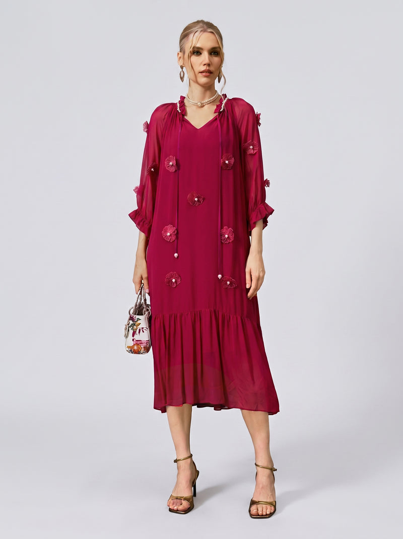 A woman wearing the Social Queen 3D Floral Pearl Mulberry Silk Dress—featuring a loose, midi-length silhouette in magenta with 3D floral appliqués—holds a small handbag while standing against a light gray background.