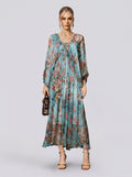 A woman stands against a plain background, wearing the Social Queen Botanical Bird Print Silk Dress—a long, flowy, A-line maxi dress with a striking blue botanical print and long sleeves. She accessorizes with a small handbag and open-toe heels.