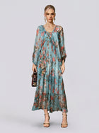 A woman stands against a plain background, wearing the Social Queen Botanical Bird Print Silk Dress—a long, flowy, A-line maxi dress with a striking blue botanical print and long sleeves. She accessorizes with a small handbag and open-toe heels.