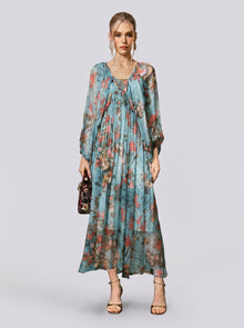 A woman stands against a plain background, wearing the Social Queen Botanical Bird Print Silk Dress—a long, flowy, A-line maxi dress with a striking blue botanical print and long sleeves. She accessorizes with a small handbag and open-toe heels.