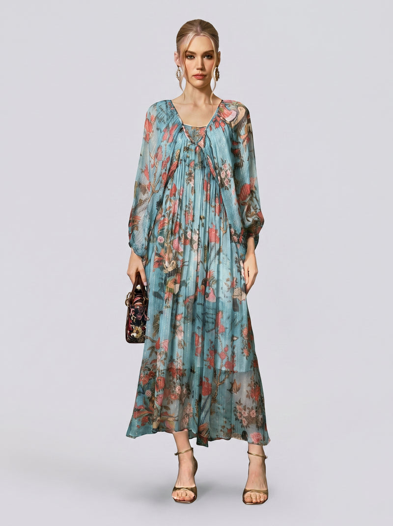 A woman stands against a plain background, wearing the Social Queen Botanical Bird Print Silk Dress—a long, flowy, A-line maxi dress with a striking blue botanical print and long sleeves. She accessorizes with a small handbag and open-toe heels.