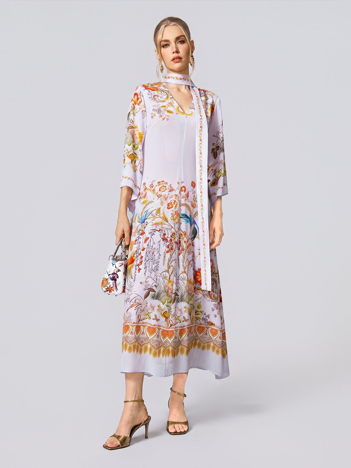 A woman wears the Social Queen Floral Bird Print Silk Dress with Scarf, featuring three-quarter sleeves and a delicate floral bird print, styled with the detachable scarf, a small purse, and beige heeled sandals against a plain background.