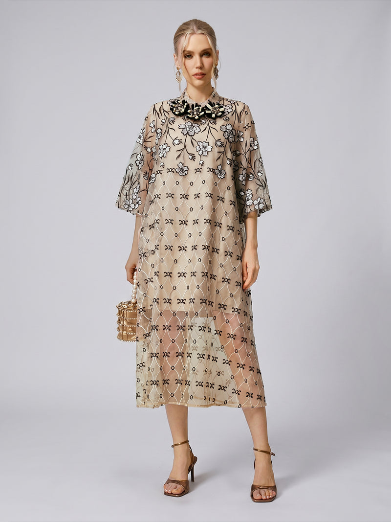 A woman stands against a plain light gray background wearing the Social Queen Floral Embroidered Mulberry Silk Dress in beige, knee-length with black detailing. She accessorizes with heeled sandals and a small bag.