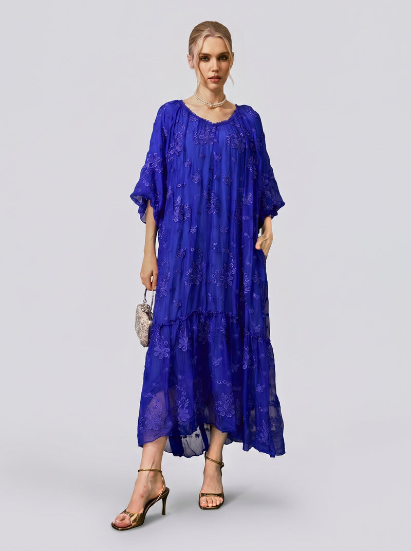 A woman stands against a plain light background, wearing the Social Queen Lace-Trim Embroidered Silk Dress—a loose-fitting, long-sleeved royal blue maxi dress featuring delicate floral embroidery and a lace-trimmed V-neckline. She accessorizes with a small purse and strappy heels.