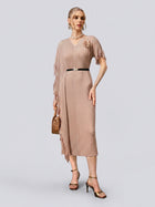 A woman wearing the Social Queen Asymmetric Fringe Midi Dress in taupe, featuring a pleated design, fringe details, and a black belt, paired with heeled sandals and a small brown handbag, stands against a plain light gray background.