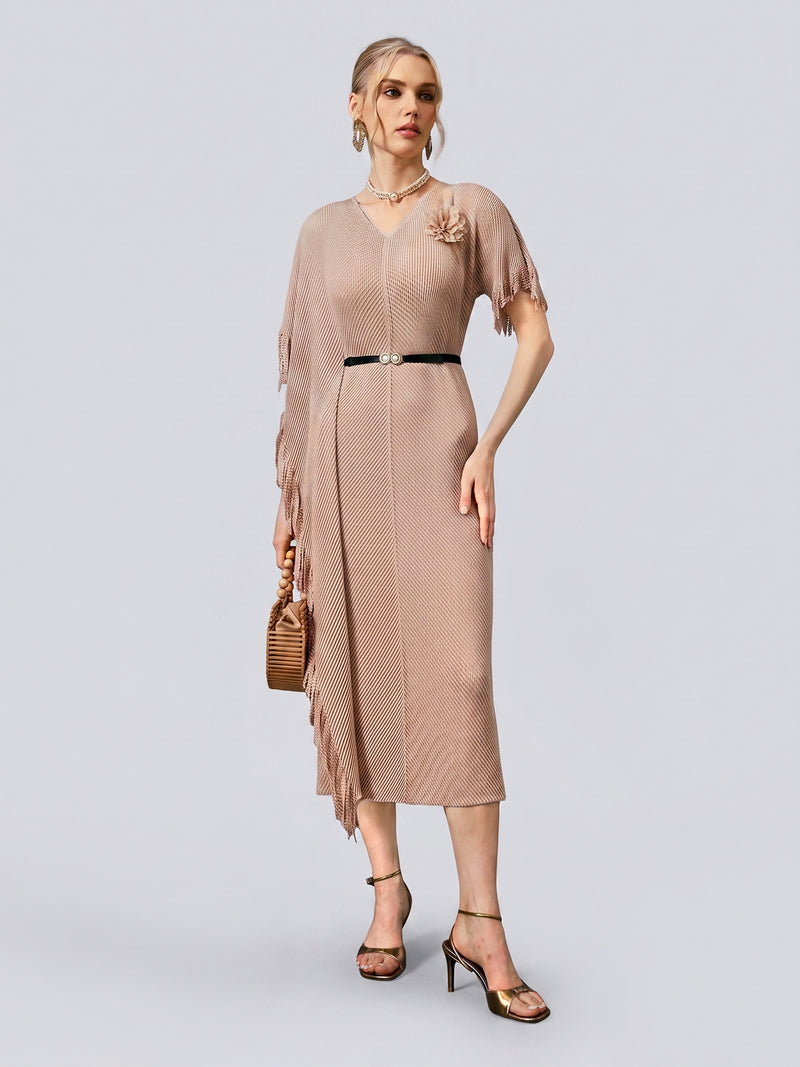 A woman wearing the Social Queen Asymmetric Fringe Midi Dress in taupe, featuring a pleated design, fringe details, and a black belt, paired with heeled sandals and a small brown handbag, stands against a plain light gray background.