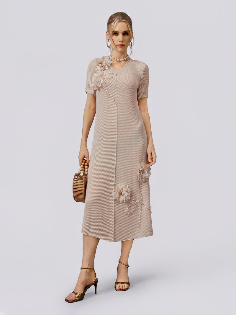 A woman stands against a plain light background wearing the Social Queen 3D Floral Pearl Embellished Pleated Dress in beige, styled with hoop earrings, heeled sandals, and a wooden handbag.