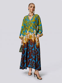 A woman wearing the Social Queen Contrast Floral Print Silk Cotton Dress—a long-sleeve, V-neck maxi dress in a blue, yellow, and burgundy floral pattern—paired with a green handbag and sandals.