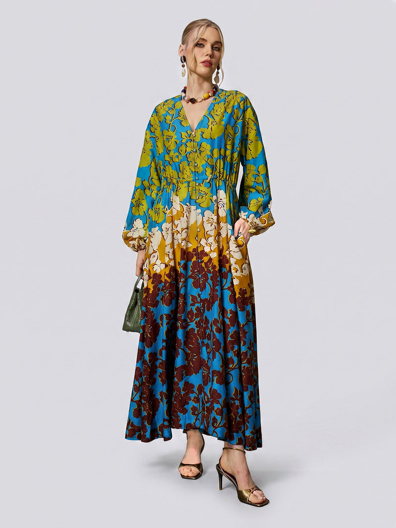 A woman wearing the Social Queen Contrast Floral Print Silk Cotton Dress—a long-sleeve, V-neck maxi dress in a blue, yellow, and burgundy floral pattern—paired with a green handbag and sandals.