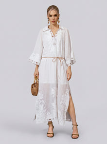 A woman stands against a plain light gray background wearing the Social Queen Floral Cutwork Embroidered Linen Dress—a long, white linen maxi dress featuring floral cutwork embroidery and a tie belt—paired with sandals, statement earrings, and a woven handbag.