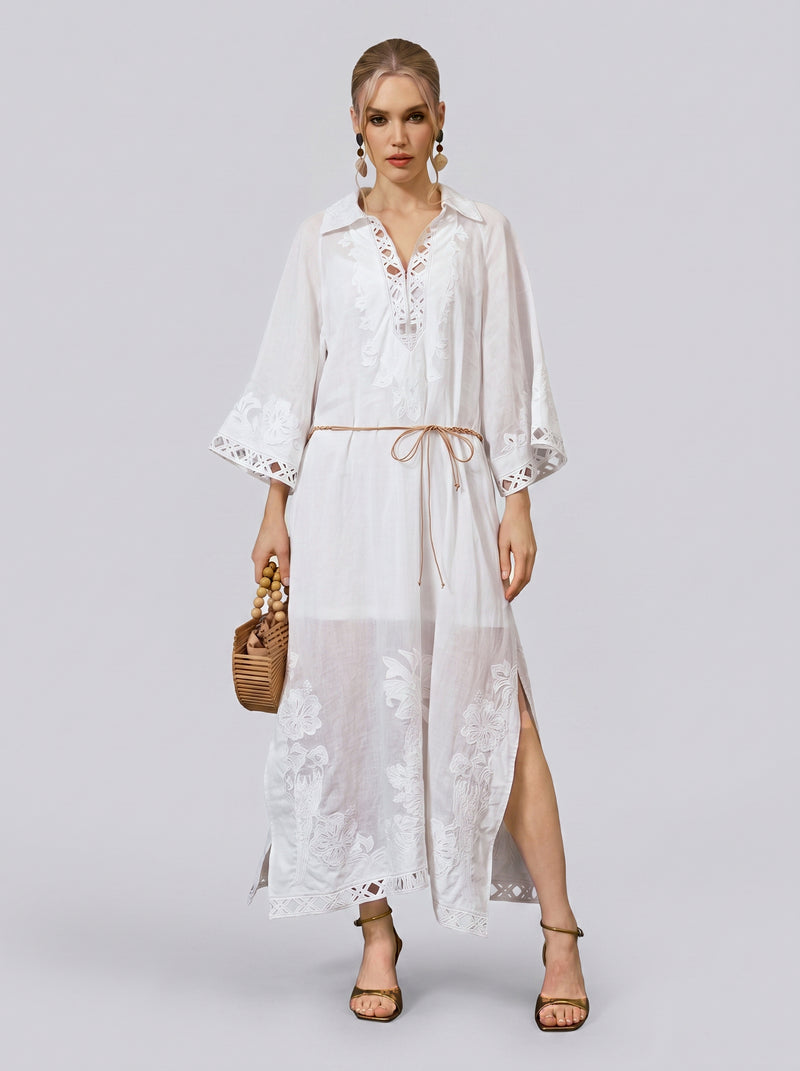 A woman stands against a plain light gray background wearing the Social Queen Floral Cutwork Embroidered Linen Dress—a long, white linen maxi dress featuring floral cutwork embroidery and a tie belt—paired with sandals, statement earrings, and a woven handbag.