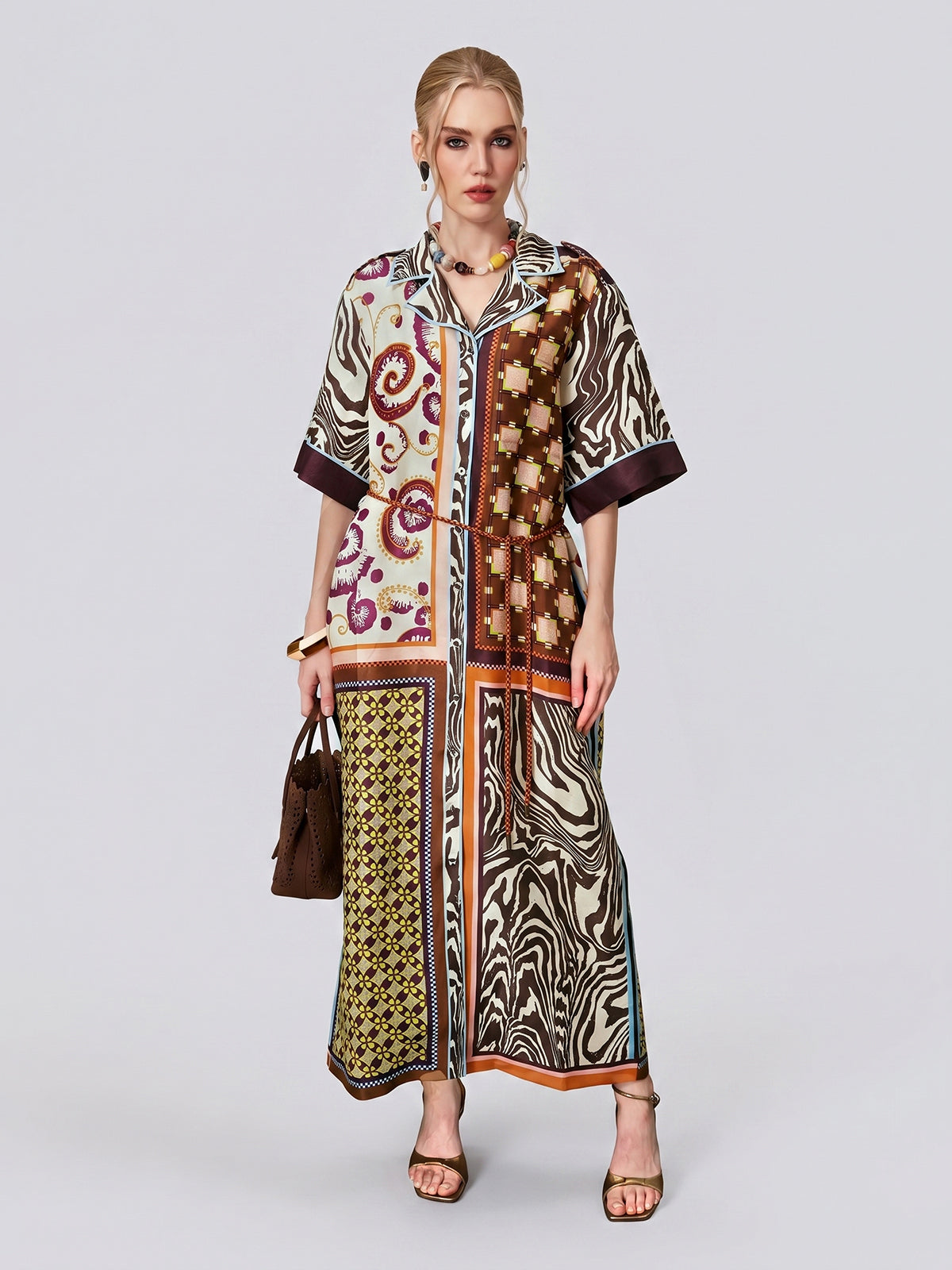 A woman wears the Social Queen Patchwork Print Cotton Shirt Dress, a loose, ankle-length dress featuring vintage patchwork and paisley motifs, styled with the included belt. She accessorizes with a brown handbag and heeled sandals while standing against a plain background.