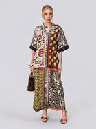 A woman wears the Social Queen Patchwork Print Cotton Shirt Dress, a loose, ankle-length dress featuring vintage patchwork and paisley motifs, styled with the included belt. She accessorizes with a brown handbag and heeled sandals while standing against a plain background.
