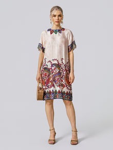 A woman stands facing forward against a plain light background, wearing the Paisley Garden Print Jacquard Silk Dress by Social Queen, which features vibrant paisley patterns with short sleeves. She accessorizes with a small handbag and heeled sandals.