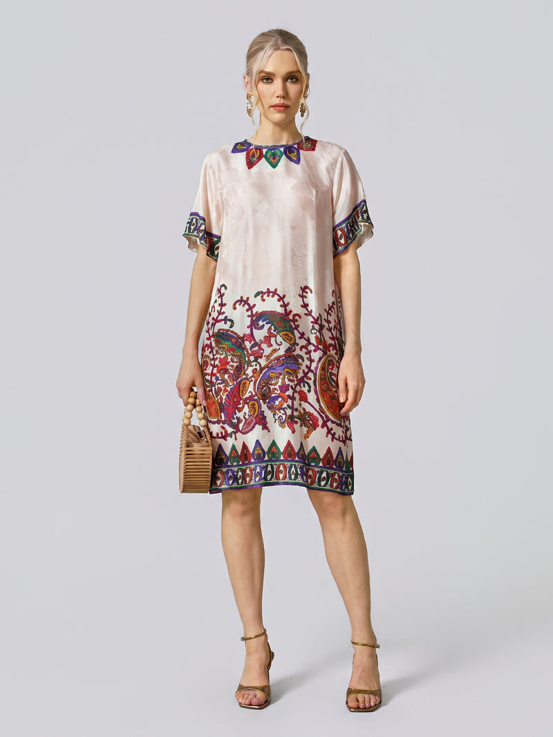 A woman stands facing forward against a plain light background, wearing the Paisley Garden Print Jacquard Silk Dress by Social Queen, which features vibrant paisley patterns with short sleeves. She accessorizes with a small handbag and heeled sandals.