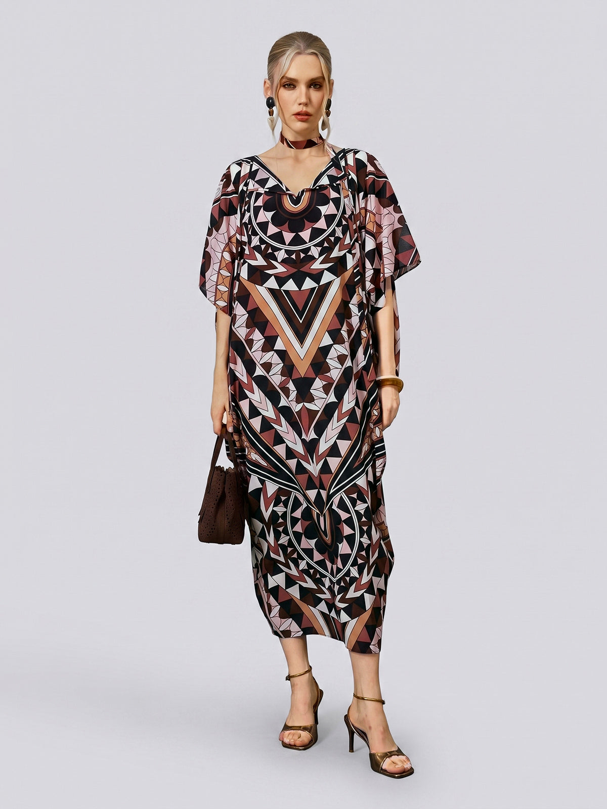 A woman stands against a plain background wearing the Social Queen Geometric Motif Print Silk Dress—with its loose fit, batwing sleeves, and black, brown, and pink geometric patterns—paired with black heels and a brown handbag. The look is completed with a coordinating scarf included with the dress.
