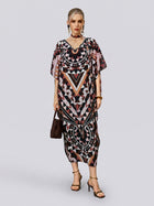 A woman stands against a plain background wearing the Social Queen Geometric Motif Print Silk Dress—with its loose fit, batwing sleeves, and black, brown, and pink geometric patterns—paired with black heels and a brown handbag. The look is completed with a coordinating scarf included with the dress.