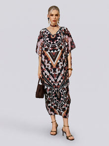 A woman stands against a plain background wearing the Social Queen Geometric Motif Print Silk Dress—with its loose fit, batwing sleeves, and black, brown, and pink geometric patterns—paired with black heels and a brown handbag. The look is completed with a coordinating scarf included with the dress.