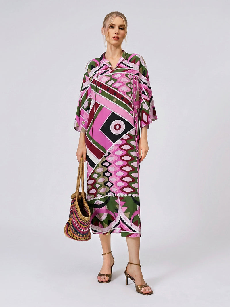 A woman wears the Social Queen Geometric Print Mulberry Silk Dress, showcasing its robe-style silhouette with pink, green, and black patterns. She accessorizes with a woven bag and heeled sandals, posed against a plain light background.