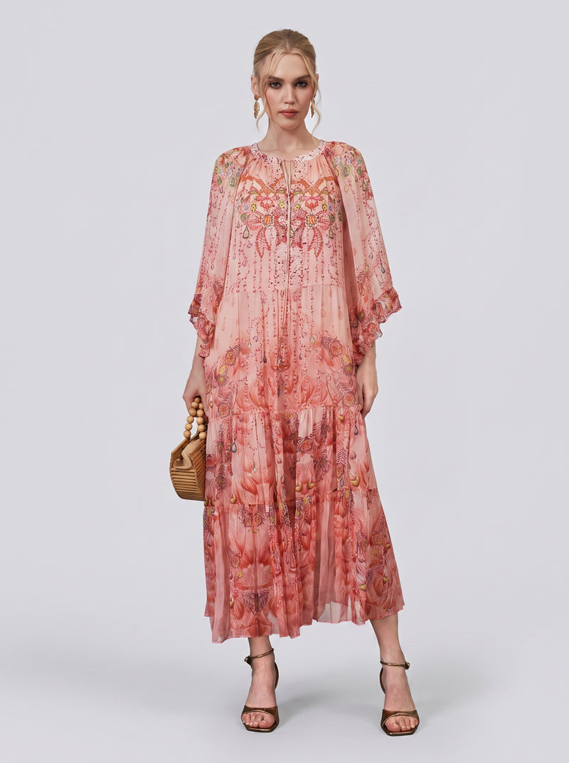 A woman stands against a plain light background, wearing the Social Queen Jewelry Print Mulberry Silk Dress—a resort-inspired maxi dress with sheer sleeves—paired with a wicker handbag and black strappy heels.
