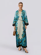 A woman stands against a plain light background wearing the Social Queen Paisley Print Linen Dress (belt included), featuring a long, robe-style silhouette in turquoise and black with a subtle paisley border print. She completes the look with a woven handbag, sandals, and a necklace.