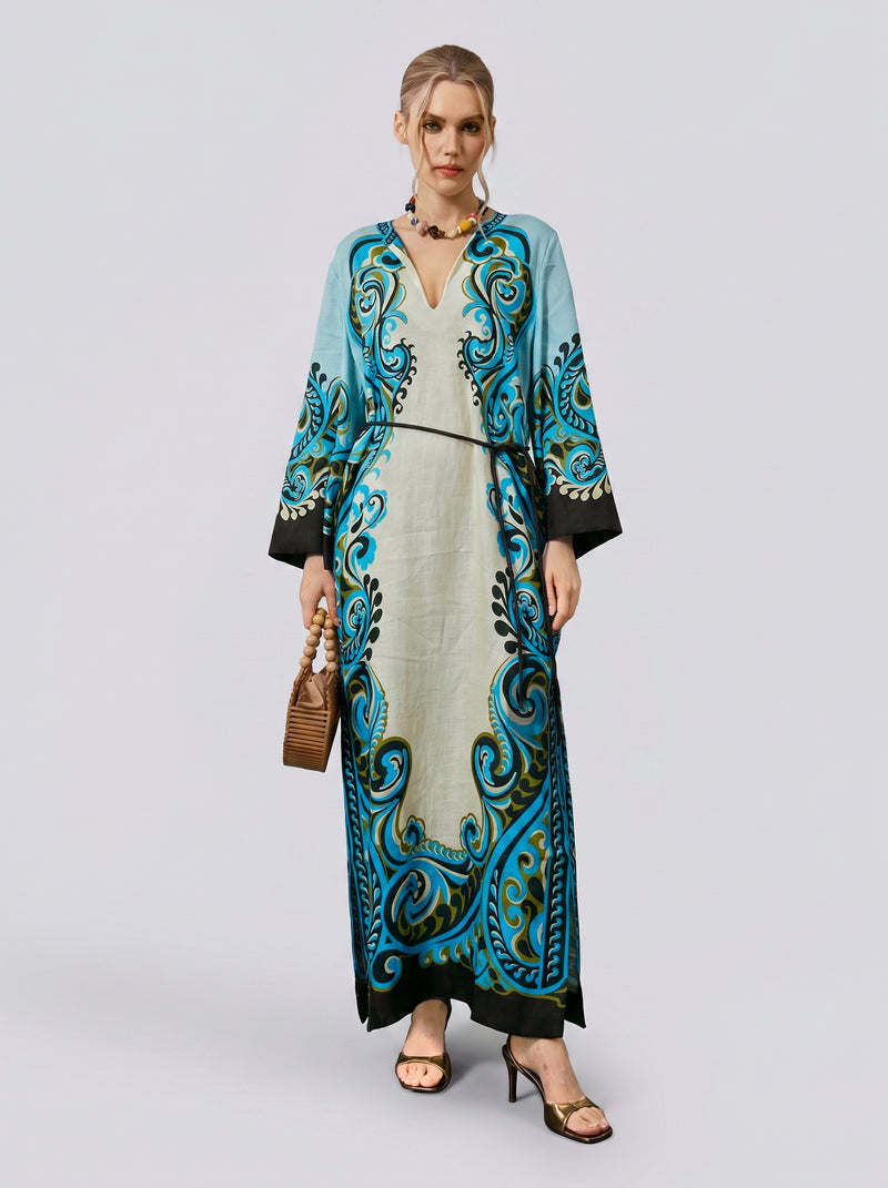 A woman stands against a plain light background wearing the Social Queen Paisley Print Linen Dress (belt included), featuring a long, robe-style silhouette in turquoise and black with a subtle paisley border print. She completes the look with a woven handbag, sandals, and a necklace.