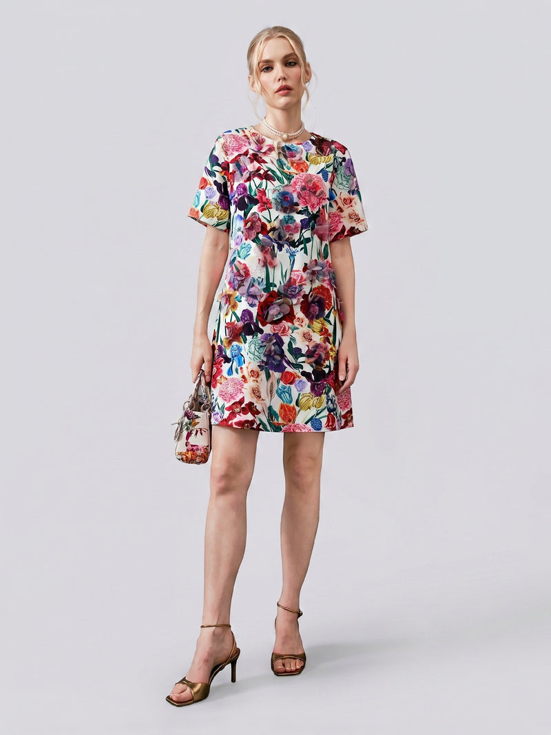 A woman stands against a plain background wearing the Social Queen 3D Floral Print Shift Dress, featuring delicate 3D organza appliqué on its short sleeves. She accessorizes with a matching floral handbag and strappy heels.