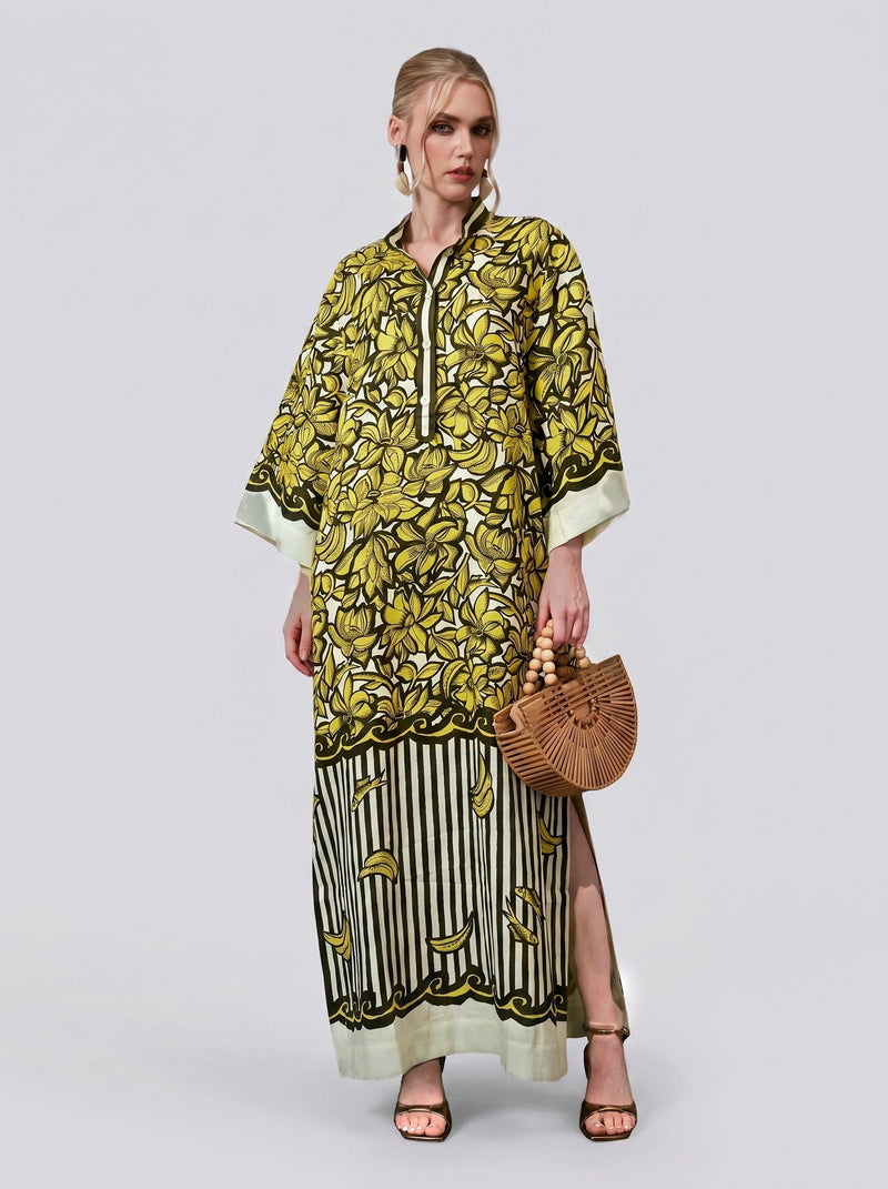 A woman stands against a plain light background wearing the Social Queen Tropical Fruit Print Linen Dress—a long dress with a yellow and black floral-inspired pattern and a stylish slit, perfect for resort wear—paired with a wooden handbag and black sandals.