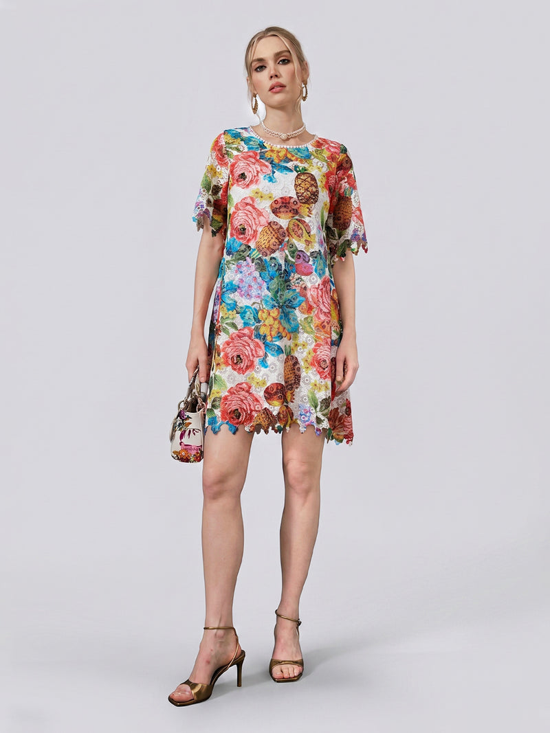 A woman models the Social Queen Botanical Fruit Print Lace Dress, a short-sleeve, knee-length resort mini dress featuring a vibrant fruit and floral print. She pairs it with heels and a coordinating floral handbag, standing against a plain light backdrop.