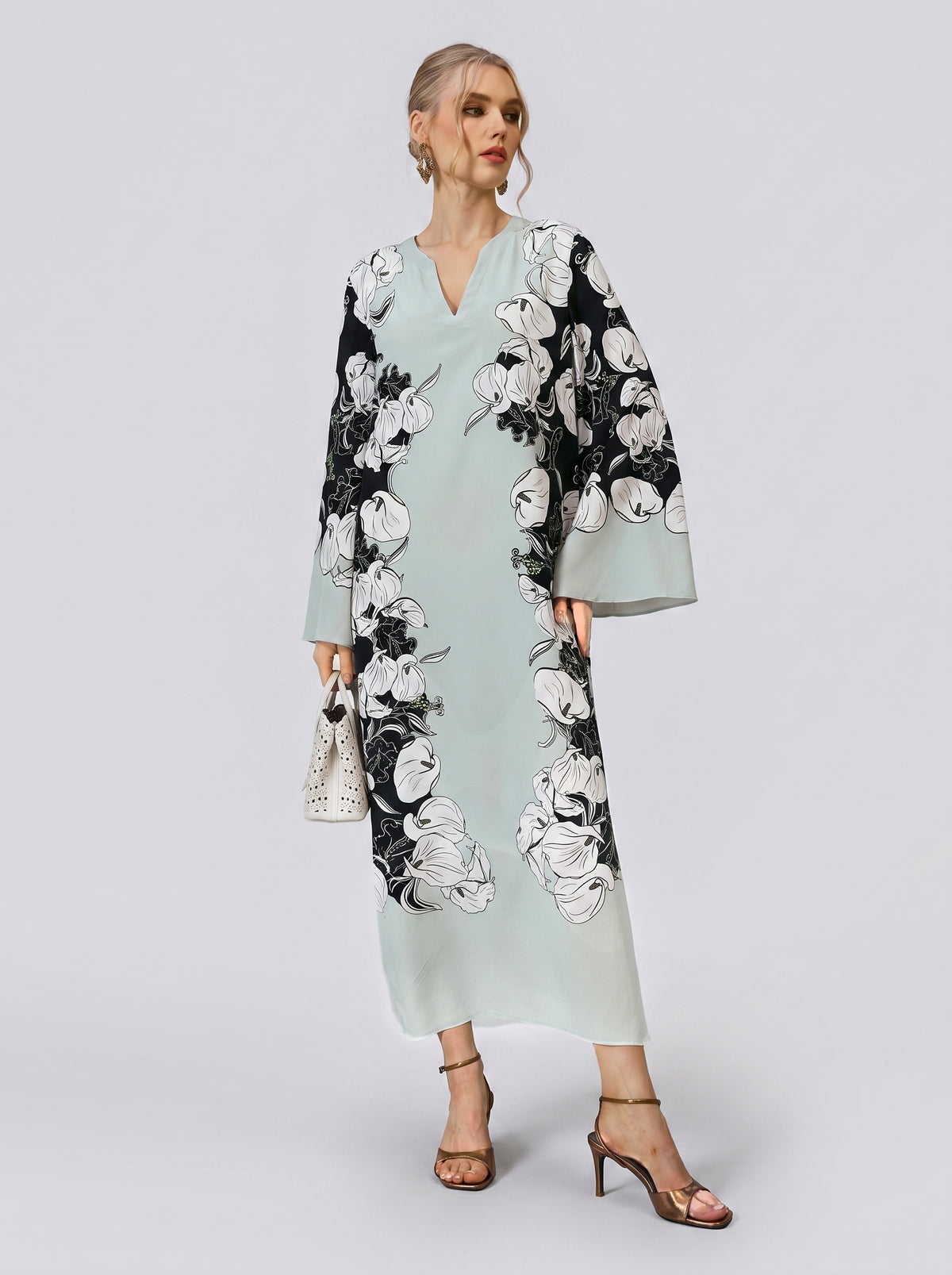A woman wearing the Social Queen Calla Lily Print Silk Dress—a long, light blue mulberry silk dress with a black and white calla lily print—paired with a white handbag and heeled sandals, stands against a plain background, making it the perfect sophisticated resort wear choice.