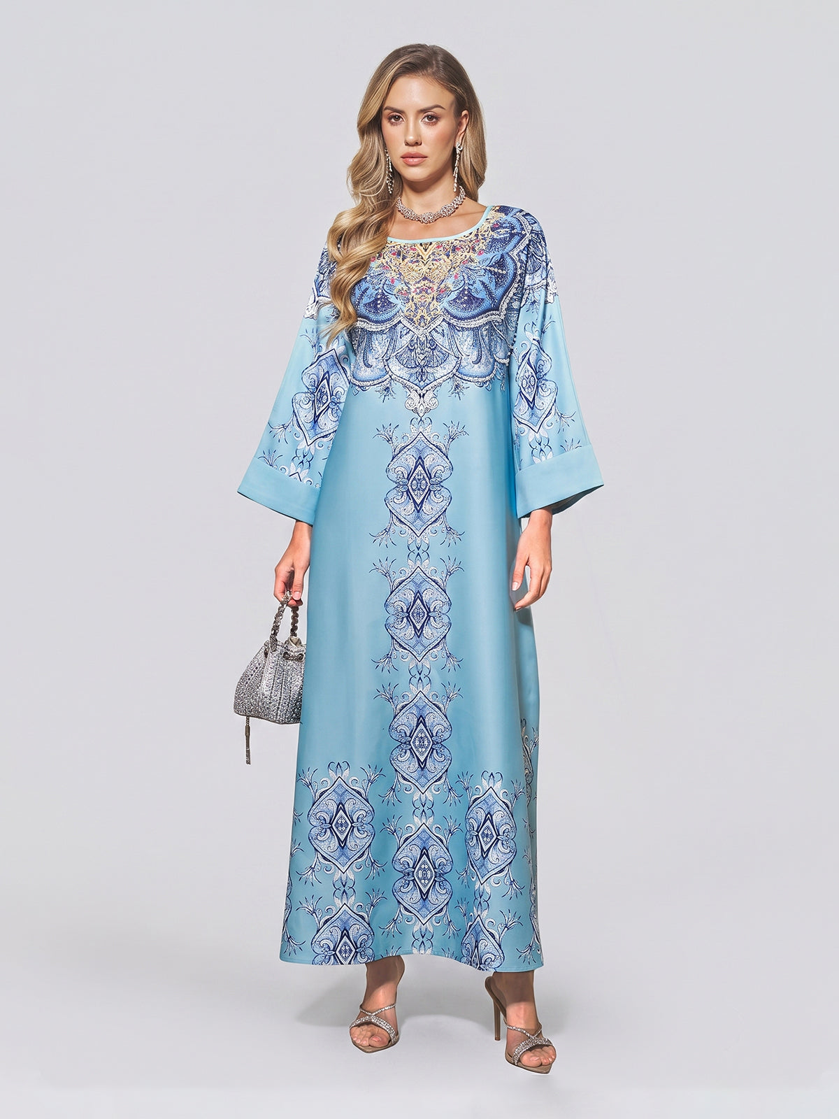 A woman wearing the Social Queen Beaded Baroque A-Line Dress—a light blue, long-sleeved, floor-length kaftan adorned with intricate blue and white beaded patterns—paired with a small silver handbag and silver sandals against a plain background.