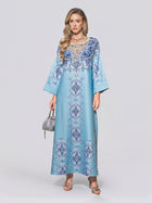 A woman wearing the Social Queen Beaded Baroque A-Line Dress—a light blue, long-sleeved, floor-length kaftan adorned with intricate blue and white beaded patterns—paired with a small silver handbag and silver sandals against a plain background.