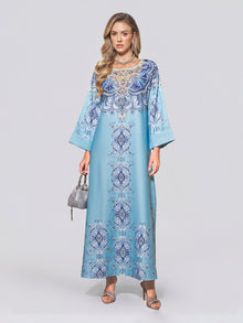 A woman wearing the Social Queen Beaded Baroque A-Line Dress—a light blue, long-sleeved, floor-length kaftan adorned with intricate blue and white beaded patterns—paired with a small silver handbag and silver sandals against a plain background.