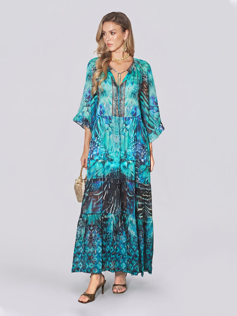 A woman wears the Beaded Floral Print Ruffled Dress by Social Queen—a long, flowing dress featuring a blue floral pattern with metallic accents and beaded embellishments. She pairs it with sandals, gold jewelry, and carries a small textured handbag while standing against a plain light background.