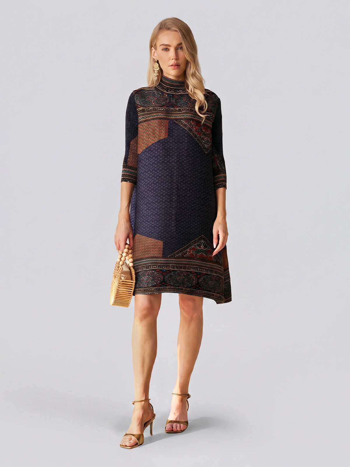 A woman stands against a plain light background wearing the Retro Ethnic Print Pleated Dress by Social Queen, featuring three-quarter sleeves and a dark jacquard satin finish. She completes her look with gold heels and a small woven handbag.