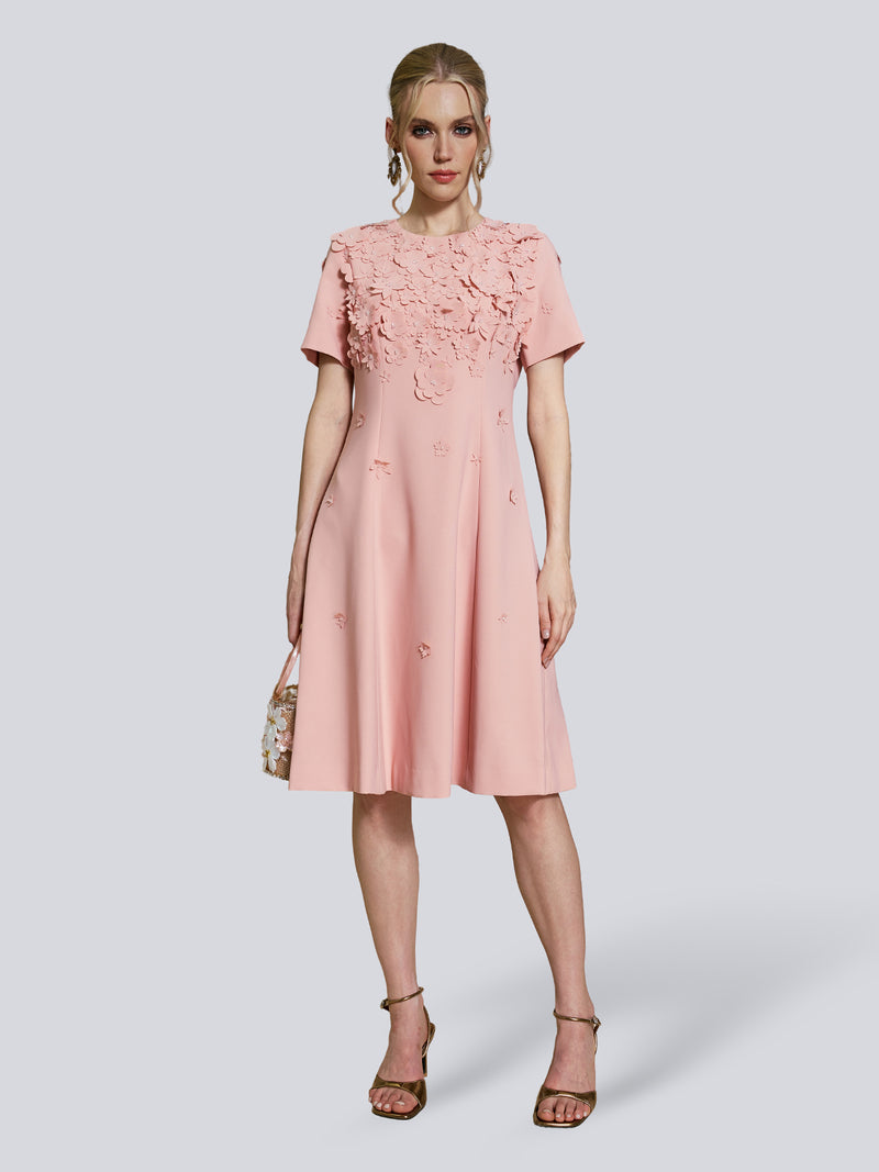 A woman is wearing the Social Queen Floral Beaded Puff Sleeve Dress, a knee-length pale pink party dress with hand-beaded floral appliqué details and short puff sleeves. She holds a small bag and stands in strappy heeled sandals.
