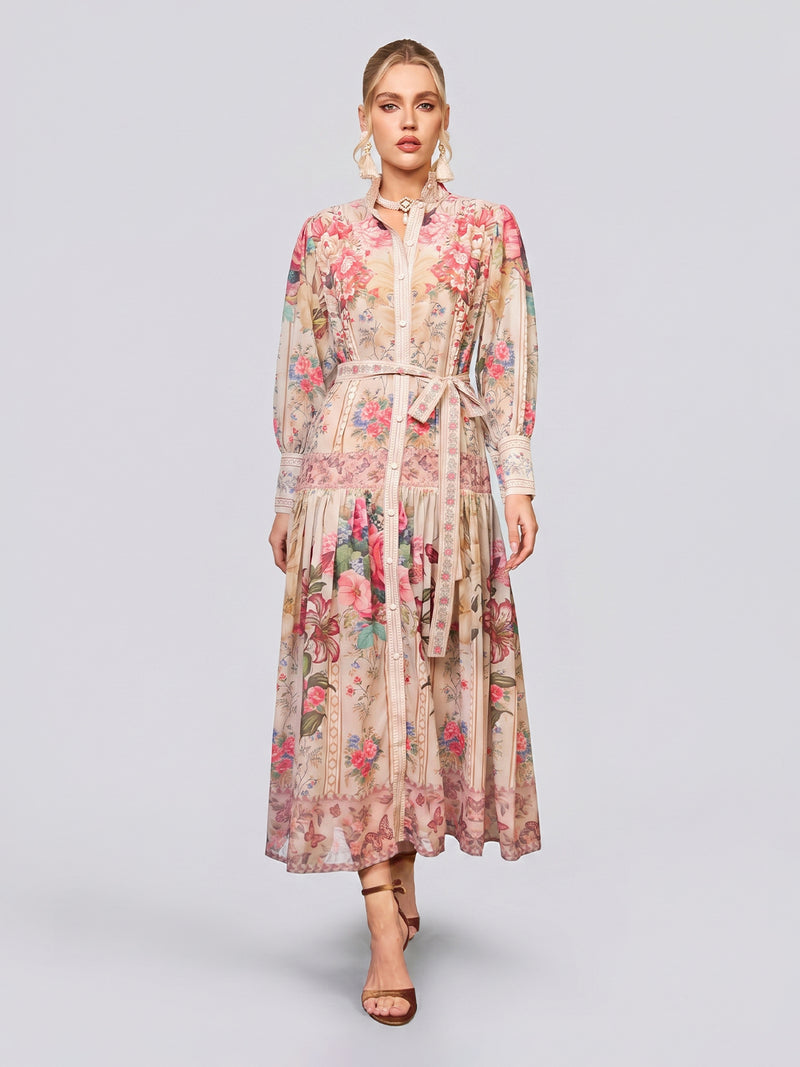 A woman is wearing the Social Queen Eden Garden Print Tie-Waist Dress—a long, sheer design with long sleeves, a buttoned front, and a flattering tie waist—paired with brown heeled sandals against a plain light background, making it the perfect choice for a stylish wedding guest look.