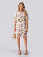 Dressed in the Social Queen Dimensional Embroidered Beaded Lace Dress, she stands against a light background, carrying a small woven handbag and wearing heeled sandals—a perfect look for a stylish wedding guest.
