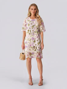 Dressed in the Social Queen Dimensional Embroidered Beaded Lace Dress, she stands against a light background, carrying a small woven handbag and wearing heeled sandals—a perfect look for a stylish wedding guest.