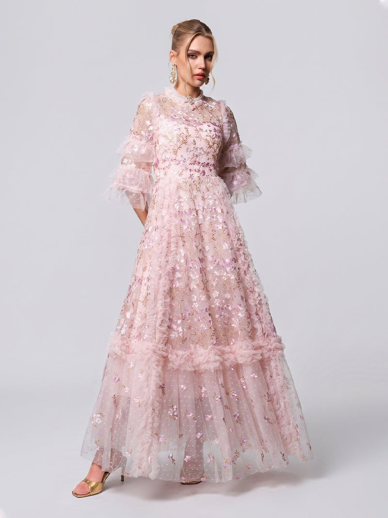 A woman wears the Social Queen Floral Embroidered Layered Tulle Dress, featuring sheer sleeves and ruffle details in a long, light pink design—an elegant choice for a wedding guest look against a simple, light background.