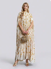 A woman stands against a plain background, dressed in the Social Queen Hand-Embellished Floral Caftan Dress featuring cream fabric adorned with gold floral embroidery. She accessorizes her look with a small beaded bag and gold sandals—creating an effortlessly elegant ensemble ideal for resort wear.