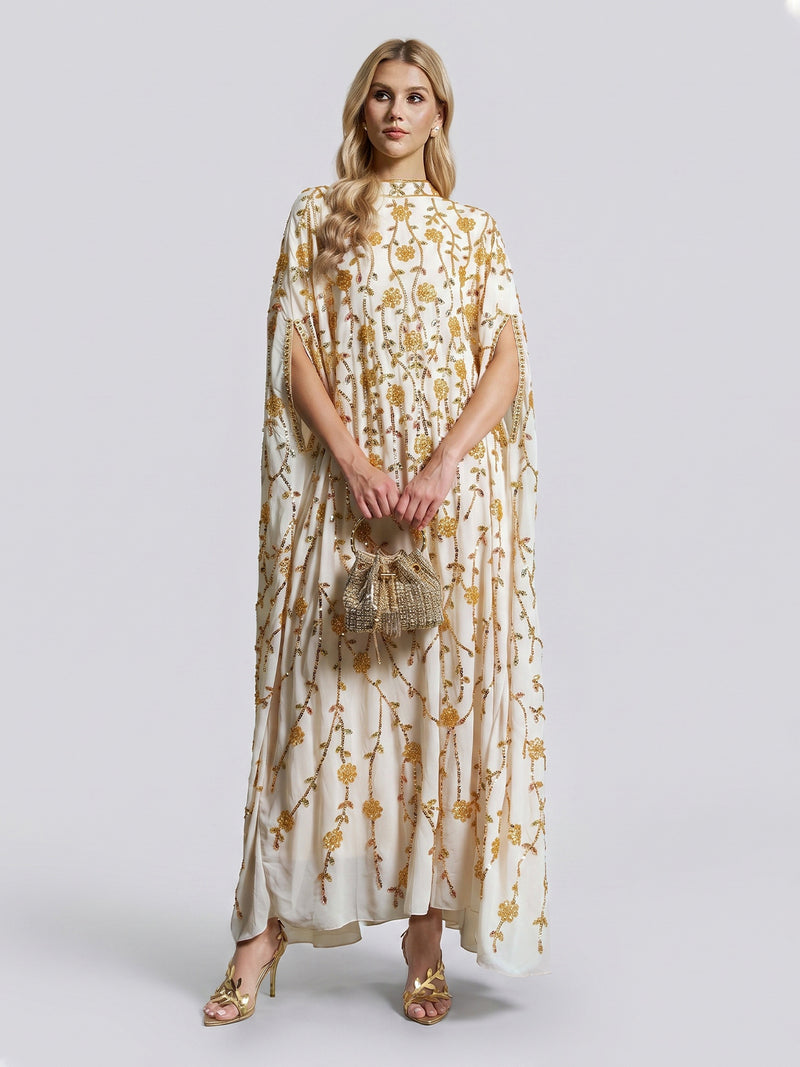 A woman stands against a plain background, dressed in the Social Queen Hand-Embellished Floral Caftan Dress featuring cream fabric adorned with gold floral embroidery. She accessorizes her look with a small beaded bag and gold sandals—creating an effortlessly elegant ensemble ideal for resort wear.