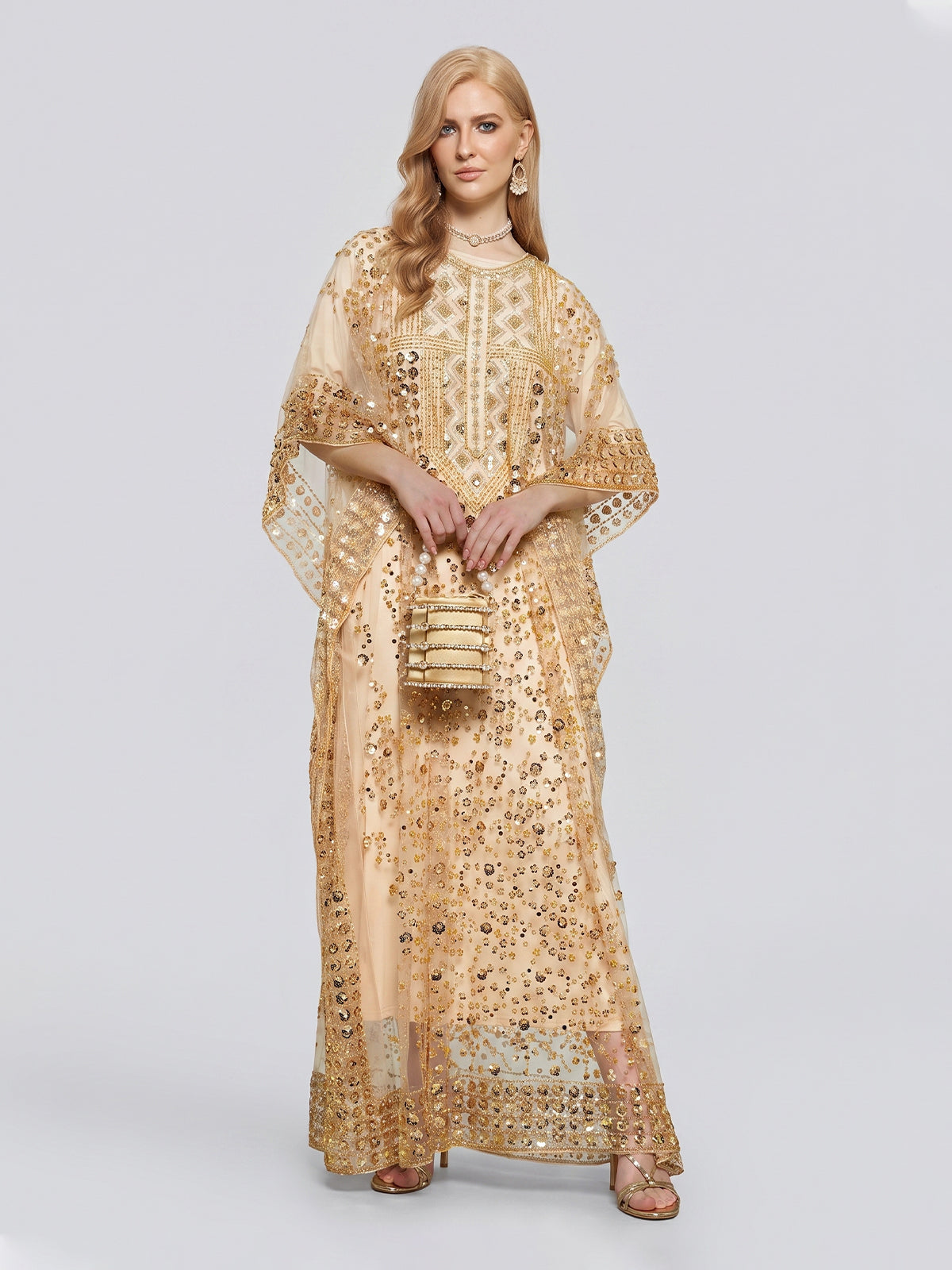 A woman wearing the Social Queen Gold Sequin Beaded Tulle Cape Dress holds a matching purse while standing against a plain light background.