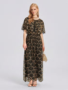 A woman stands against a plain light background wearing the Social Queen Gilded Beaded Cape Dress, styled with gold sandals, coordinating jewelry, and a small gold handbag.