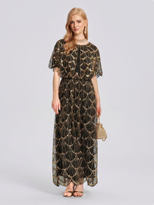 A woman stands against a plain light background wearing the Social Queen Gilded Beaded Cape Dress, styled with gold sandals, coordinating jewelry, and a small gold handbag.
