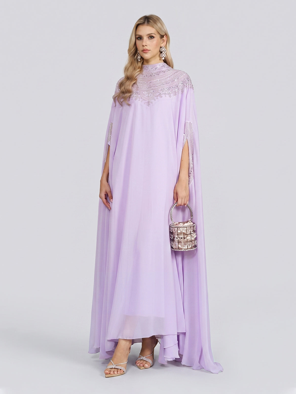 A woman wearing the Social Queen Beaded Tulle Caftan Cape Dress with delicate lace details stands against a plain background, holding a small handbag and wearing heeled sandals.